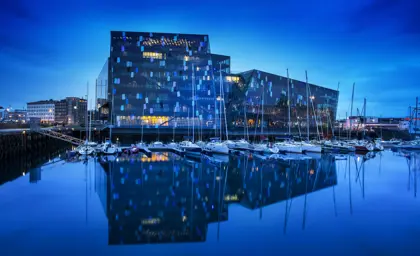 Harpa Concert Hall in Reykjavík, Iceland, at night with a view of its glass facade reflecting on the calm waters of the harbor with sailboats moored nearby.