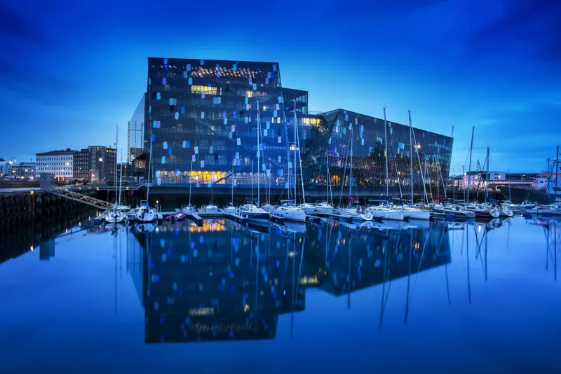 Harpa Concert Hall in Reykjavík, Iceland, at night with a view of its glass facade reflecting on the calm waters of the harbor with sailboats moored nearby.