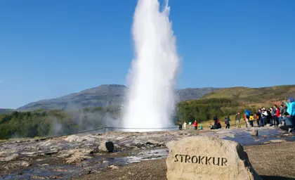 Strokkur geyser erupting under a clear blue sky with tourists watching from a safe distance in Geysir geothermal area, Iceland