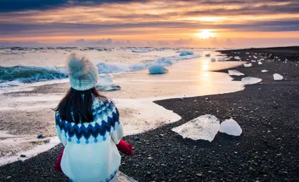 Woman wearing Icelandic knit sweater sitting on ice chunk at Diamond Beach in Iceland, admiring vibrant winter sunset.