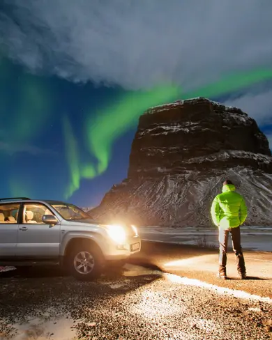 Man hunting for the northern lights in Iceland, standing next to a jeep under green lights above a mountain.