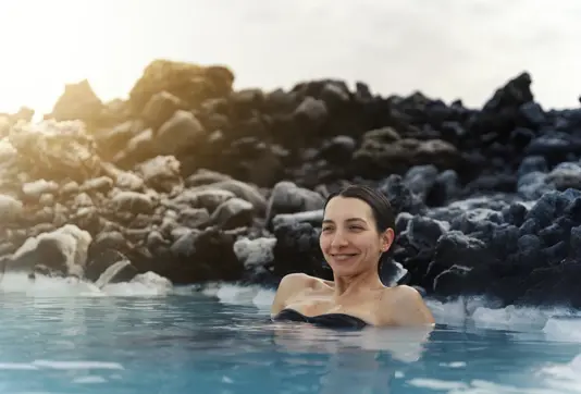 Happy woman enjoying a peaceful soak in the geothermal waters of Blue Lagoon Iceland, surrounded by black lava rocks at sunset—capturing a moment of wellness on a Travel Reykjavik spa experience.