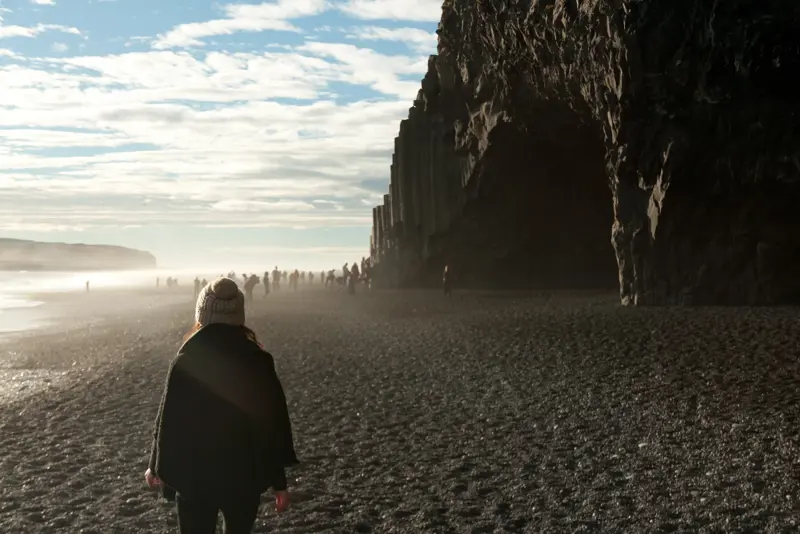 Woman walks toward the cave in Reynisfjara beach at sunrise with tourists exploring the dramatic basalt cliffs on Iceland's southern coast.